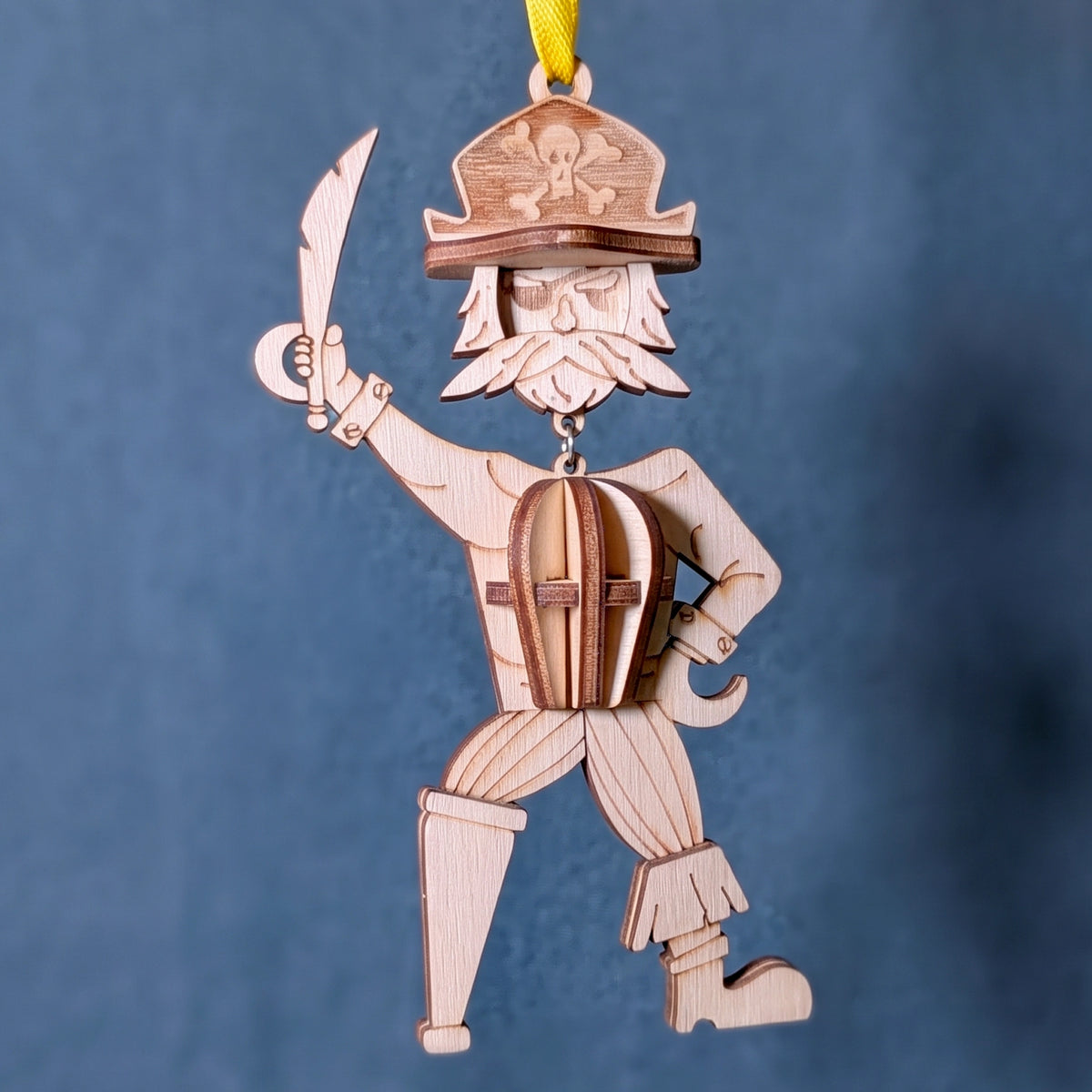 Hanging 3D laser cut plywood pirate ornament with hook hand, cutlass, and peg leg suspended by yellow ribbon on blue fabric.