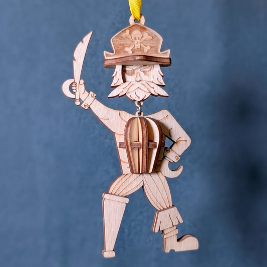 Hanging 3D laser cut plywood pirate ornament with hook hand, cutlass, and peg leg suspended by yellow ribbon on blue fabric.