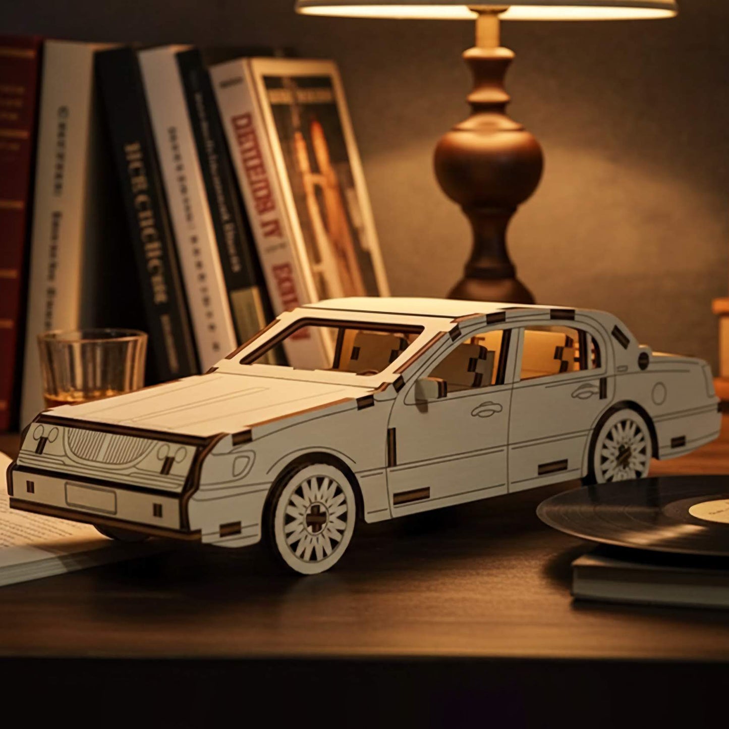 Laser cut wooden plywood luxury sedan 3D model on a wooden desk next to a vintage lamp and vinyl records. Detailed plywood assembly featuring a classic grille, multi-spoke wheels, and realistic door handles. Warm evening atmosphere with bokeh lights.