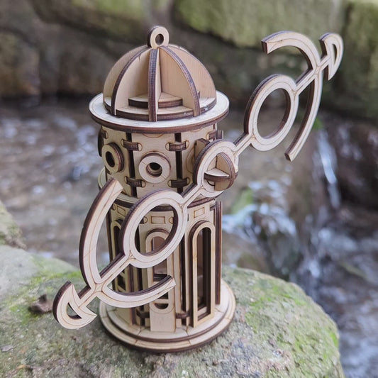Small laser cut plywood windmill tower toy with tilted anchor wings, standing on a mossy root in a blurry woods.