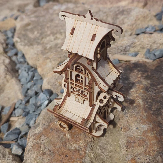 A miniature fairytale caravan model built of laser cut plywood against a blurry background of trees.
