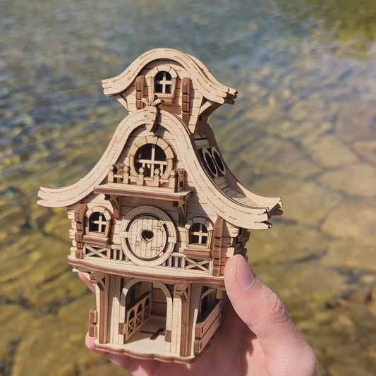 A hand holding a miniature laser cut plywood bunny house puzzle against a soft blurred bright outdoor garden background.