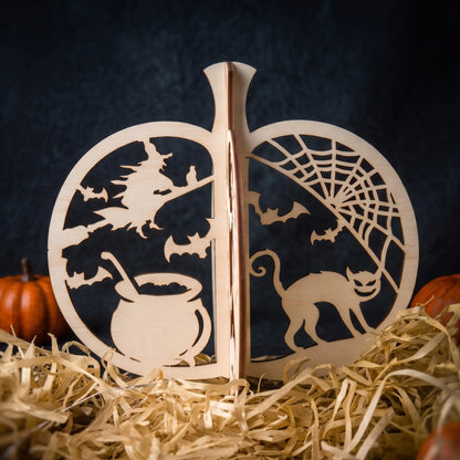 A wooden laser cut Halloween decorative pumpkin with laser-cut scenes including a witch on a broomstick, bats, a cauldron, a spiderweb, and a cat, displayed on straw with small pumpkins in the background.