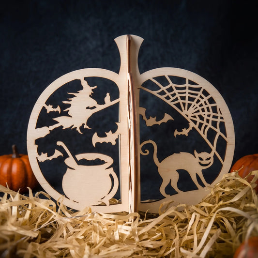 A wooden laser cut Halloween decorative pumpkin with laser-cut scenes including a witch on a broomstick, bats, a cauldron, a spiderweb, and a cat, displayed on straw with small pumpkins in the background.