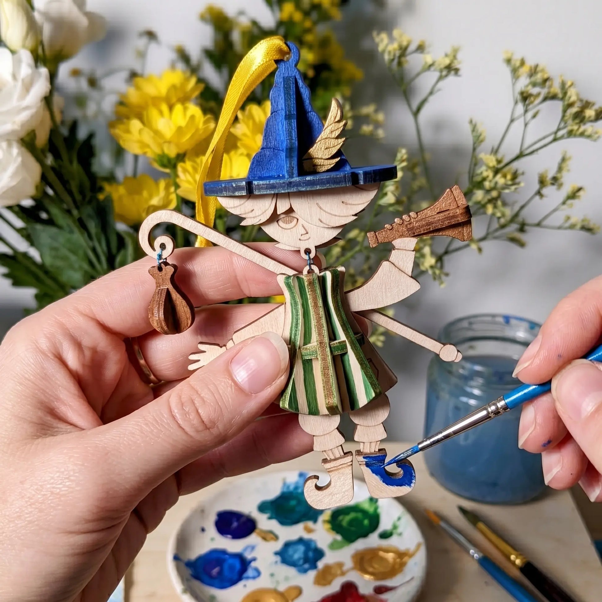 Paint your own wooden marionette puppet ornament made from a laser cut SVG file, shown with brushes, paints, flowers, and colorful bard character details.