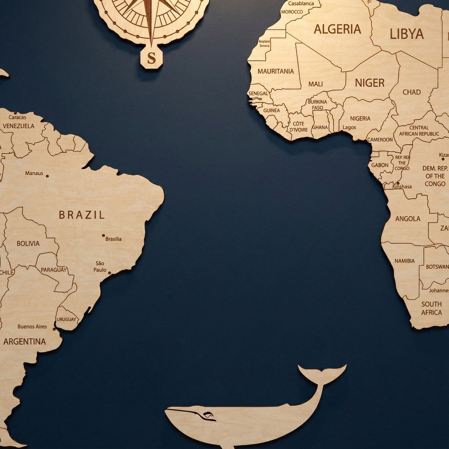 Close-up view of South America and Africa on a 3D wooden world map wall hanging. Features detailed engravings of countries like Brazil, Algeria, and South Africa, with a decorative wooden whale silhouette in the Atlantic Ocean against a dark blue background.