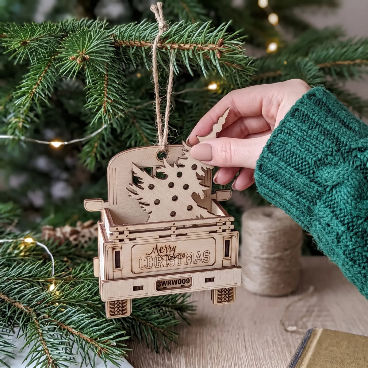 Hand inserting a wooden pine tree into a laser cut plywood vintage truck ornament hanging on fir tree branch.