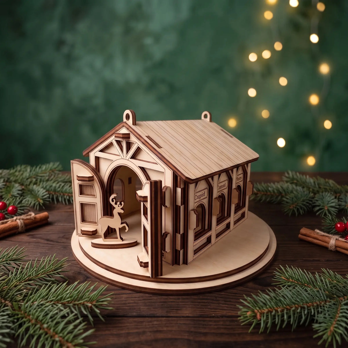 Perspective view of laser cut plywood reindeer barn on rustic table with holiday pine branches, berries, and lights.