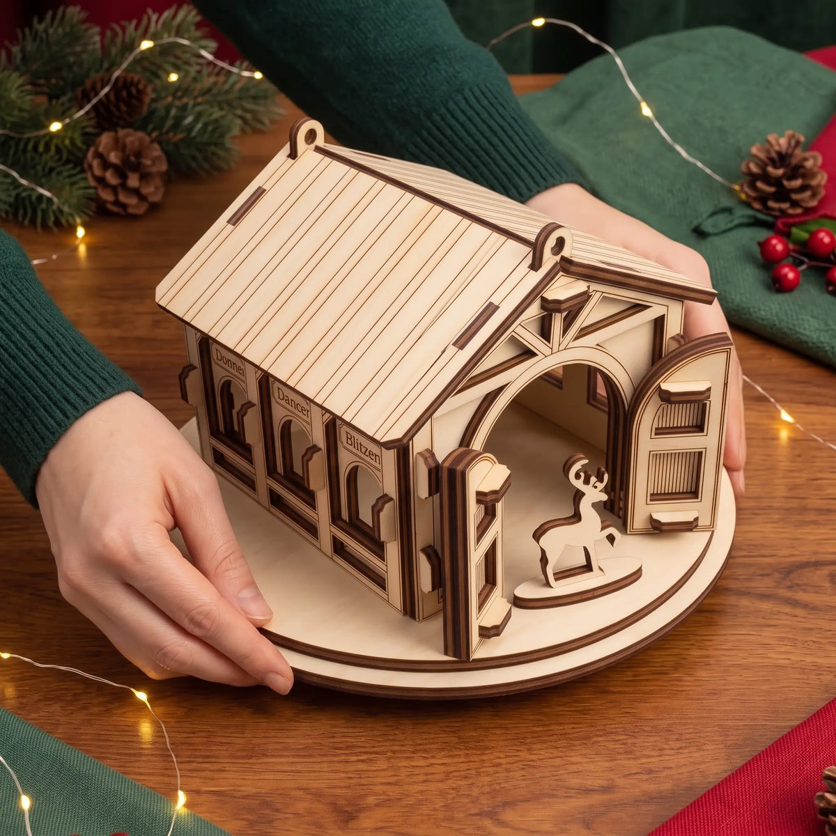 Hands display assembled laser cut plywood reindeer stable showing true scale beside festive lights and pinecones.