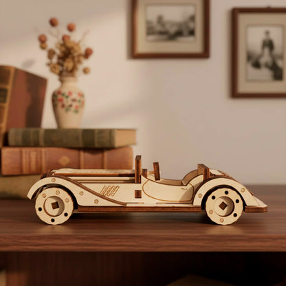 Side profile of a laser cut wooden plywood retro roadster model. Elegant plywood craft with curved fenders and open cockpit. Displayed on a bookshelf next to vintage books and a floral vase. Precision wood assembly kit with dark layered edges.