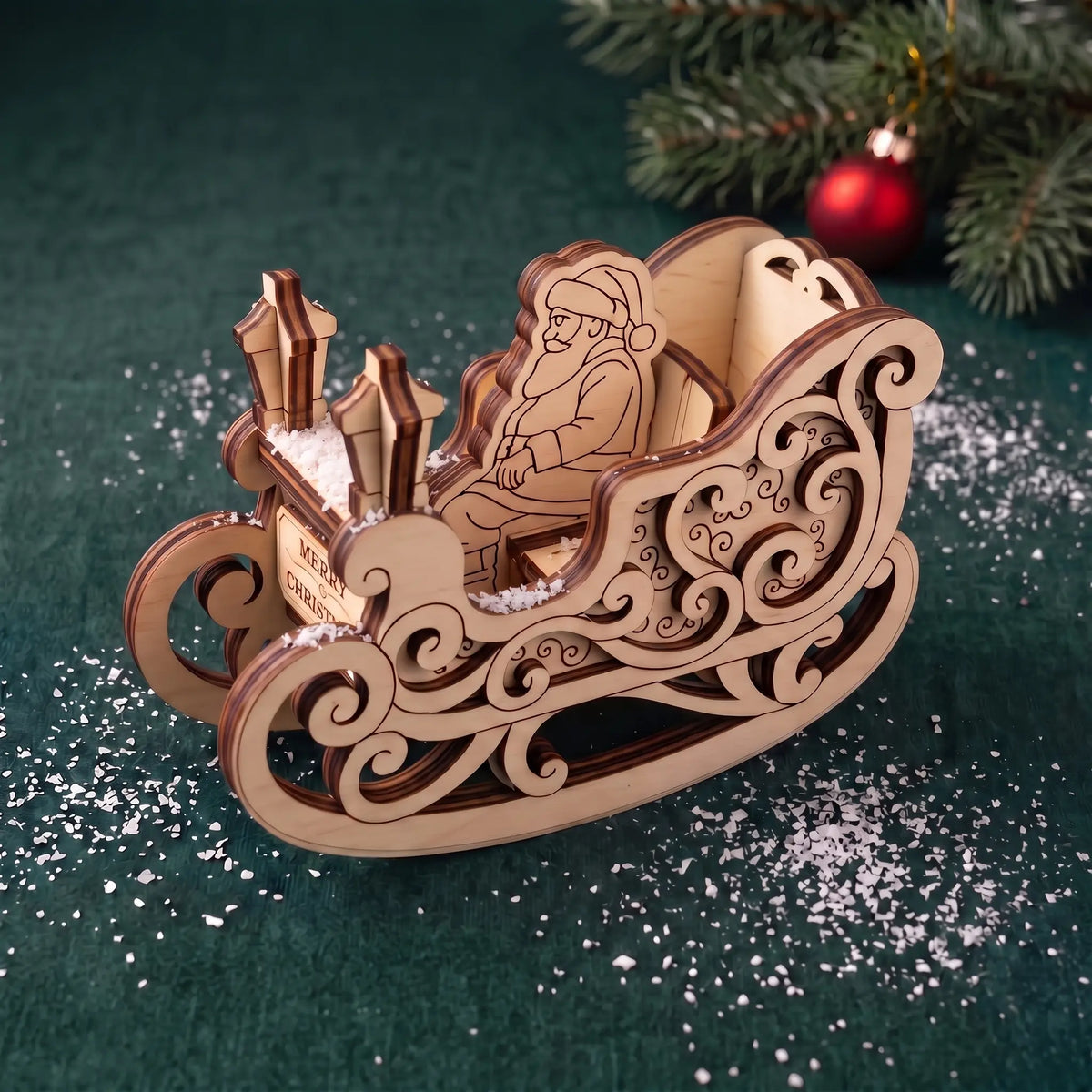 Laser cut plywood rocking sleigh with Santa figure on snowy dark green velvet by Christmas tree branches.