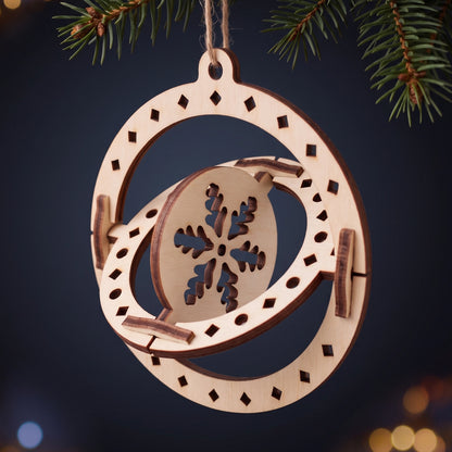 3D kinetic laser cut plywood sphere ornament with spinning snowflake hanging from fir branch with lights.