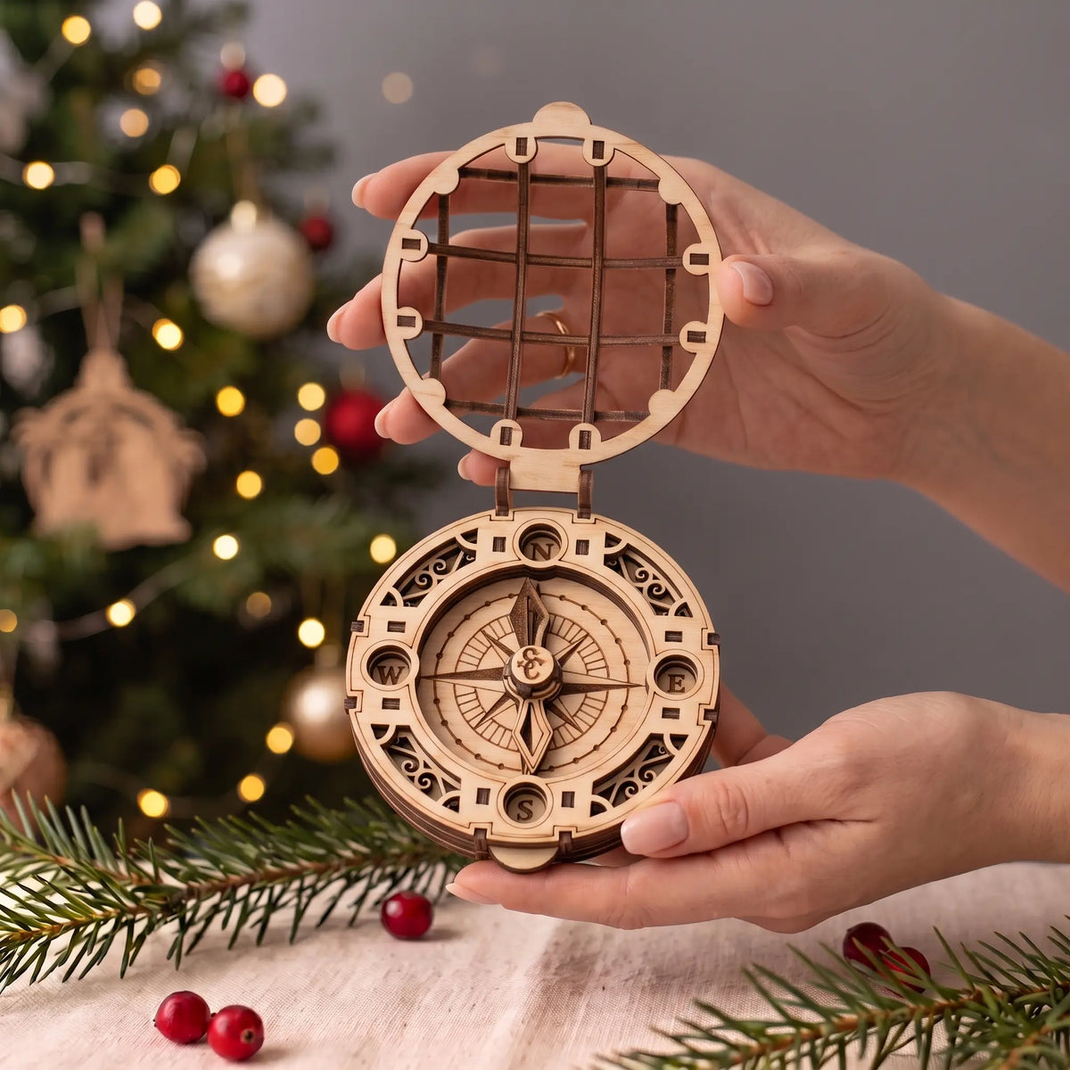 Hands hold open laser cut plywood Santa Claus compass toy showing true scale against a glowing Christmas tree.
