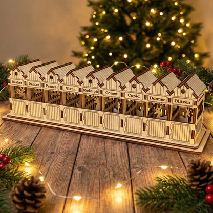 Wooden laser cut Santa's Reindeer stable with nine individual stalls named after Santa's reindeer including Rudolph, illuminated warmly against a softly blurred Christmas tree background.