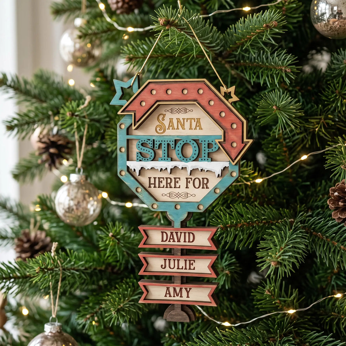 Painted laser cut plywood Santa Stop Here sign ornament with custom name plaques hanging on a bright Christmas tree.