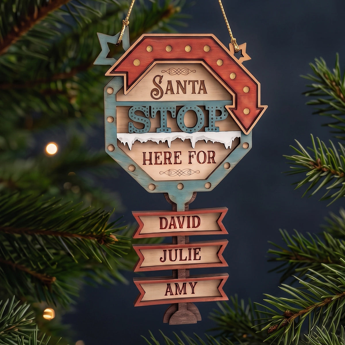 Laser cut plywood Santa Stop Here ornament with engraved name tags hanging on pine branches with glowing fairy lights.