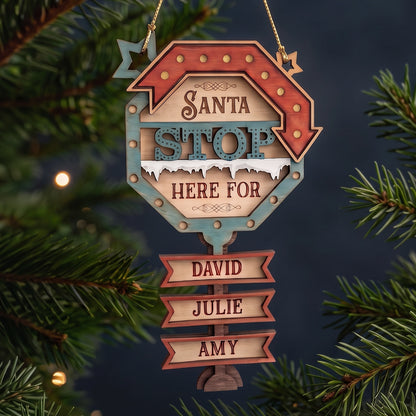 Laser cut plywood Santa Stop Here ornament with engraved name tags hanging on pine branches with glowing fairy lights.