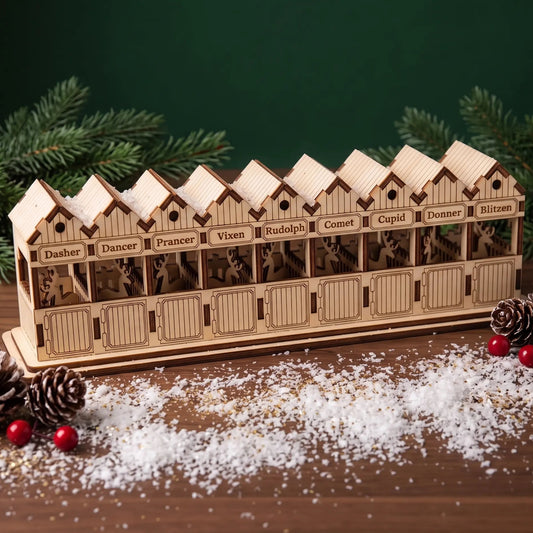 Laser cut plywood reindeer barn with snow on roofs, pinecones, and engraved names on a wooden table.