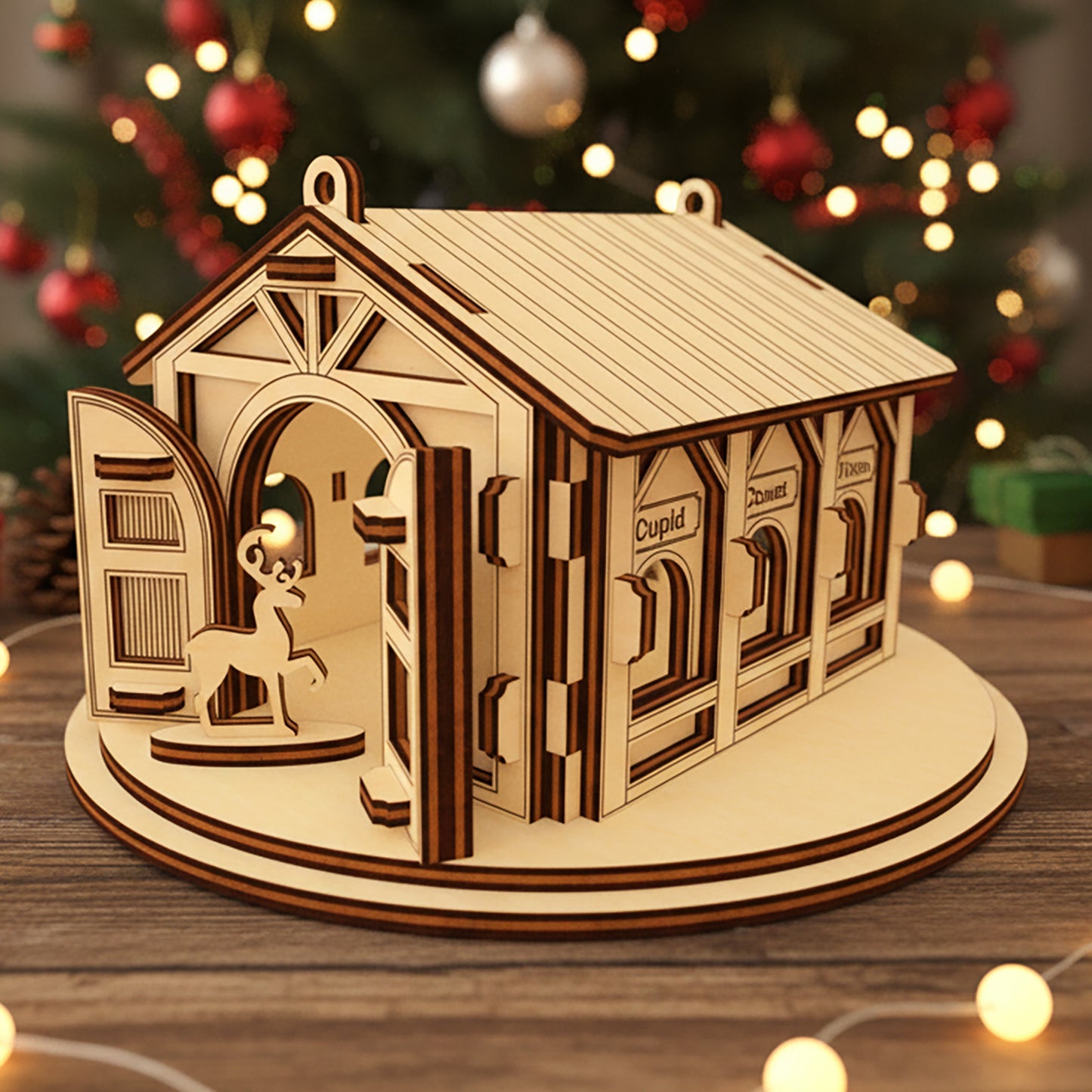 Laser cut wooden Santa's Reindeer Barn ornament with a reindeer peeking through the open door and labeled stalls for Comet, Cupid and Vixen, placed against a twinkling Christmas tree lights background.
