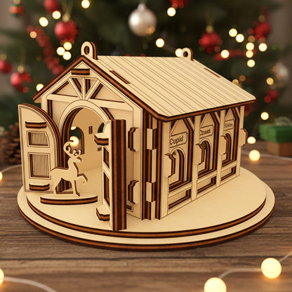 Laser cut wooden Santa's Reindeer Barn ornament with a reindeer peeking through the open door and labeled stalls for Comet, Cupid and Vixen, placed against a twinkling Christmas tree lights background.