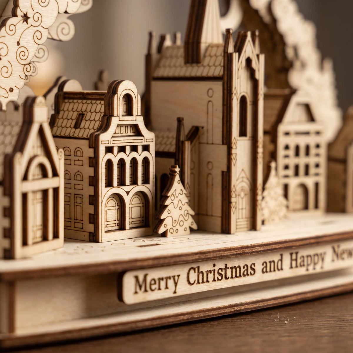 Close-up of laser cut plywood Christmas city diorama highlighting church tower, townhouse facades, and holiday lettering.