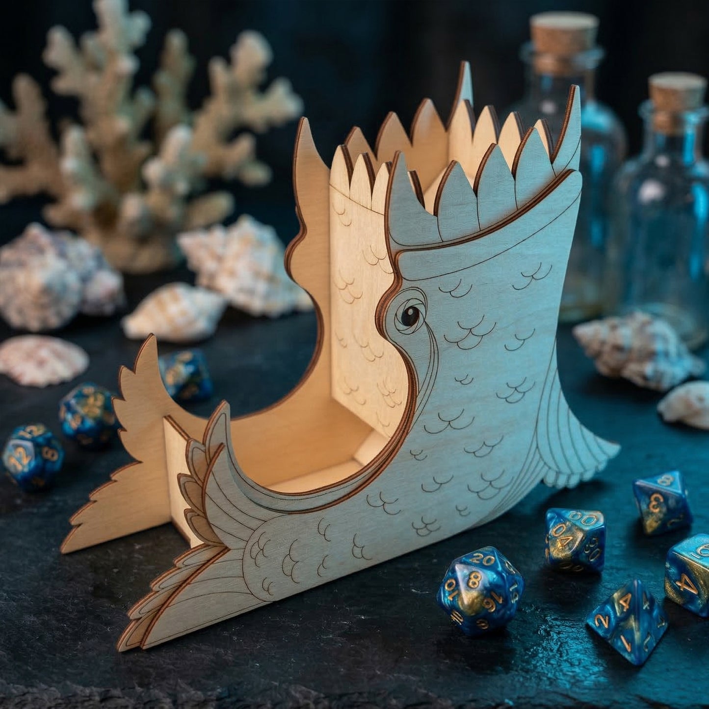 Front-angled view of a sea creature dice tower crafted from laser cut plywood. Detailed wood etching shows scales and a stylized eye. Blue RPG dice sit in the integrated landing tray on a nautical-themed table with coral and seashells.