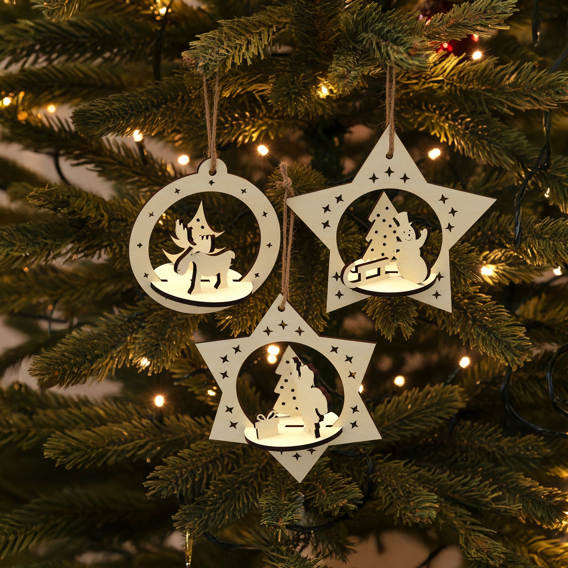 Delicate laser cut Set of 3 Christmas toys with intricate pop-out holiday figures of deer, snowman and Santa in hanging star-shaped frames, set against a twinkling Christmas tree background.