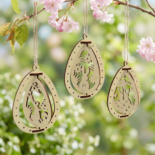 Three Easter egg ornaments made of laser cut plywood hanging from a flowering branch. Each egg features a different cutout scene with rabbits and flowers. The natural light wood grain is accented by dark brown burnt edges.