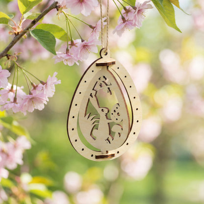 A detailed laser cut plywood Easter egg ornament featuring a whimsical rabbit reaching for a plant. The 3D layered design is suspended by a twine string against a backdrop of pink cherry blossoms in a sunny spring garden.