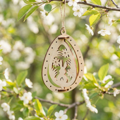 Wooden Easter egg decoration crafted from laser cut plywood with an intricate silhouette of a bunny among flowers. Hanging from a tree with white blossoms, showcasing the precise cutouts and smooth birch texture.