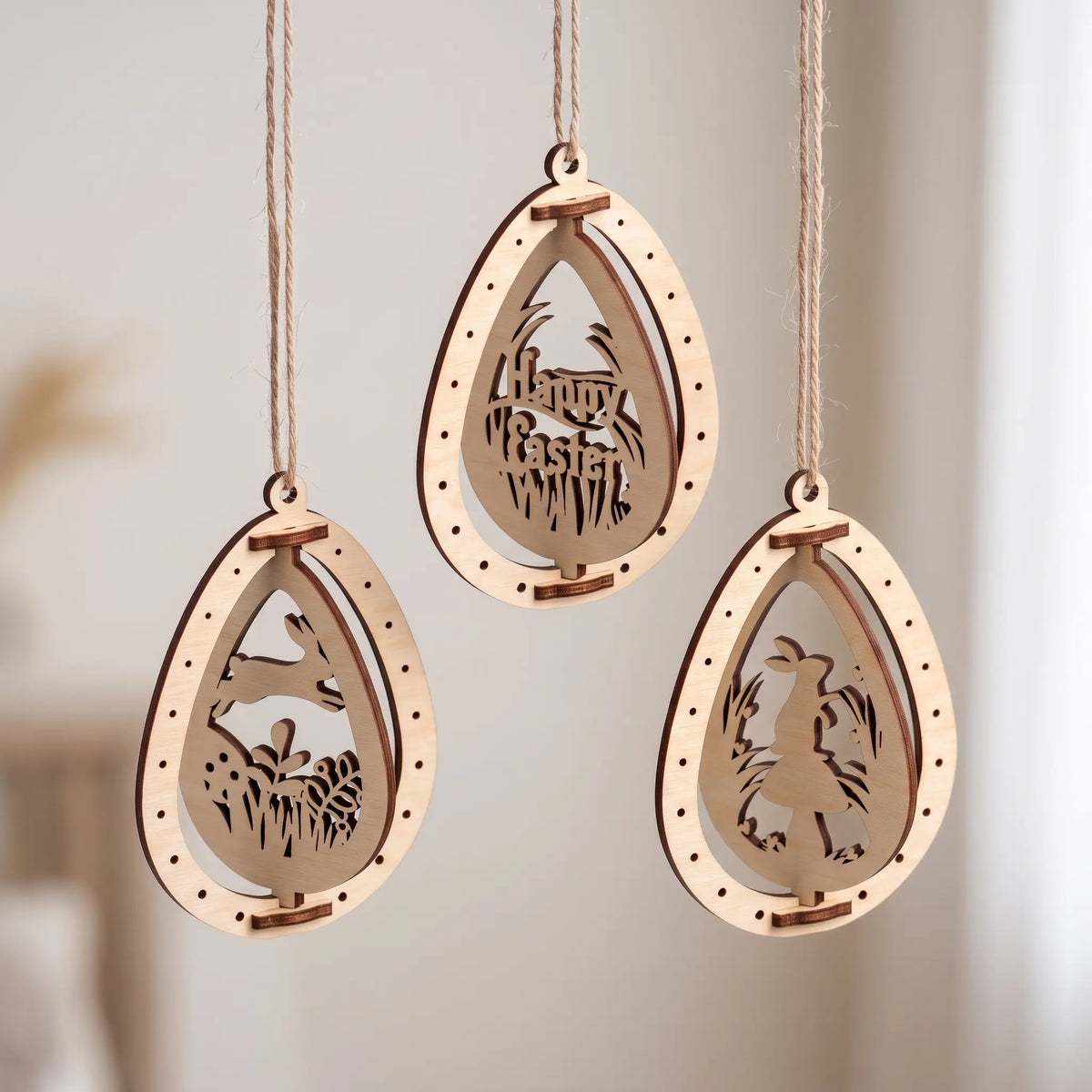 Three natural laser cut plywood Easter egg ornaments with rotating bunny and Happy Easter cutouts hanging by twine strings.