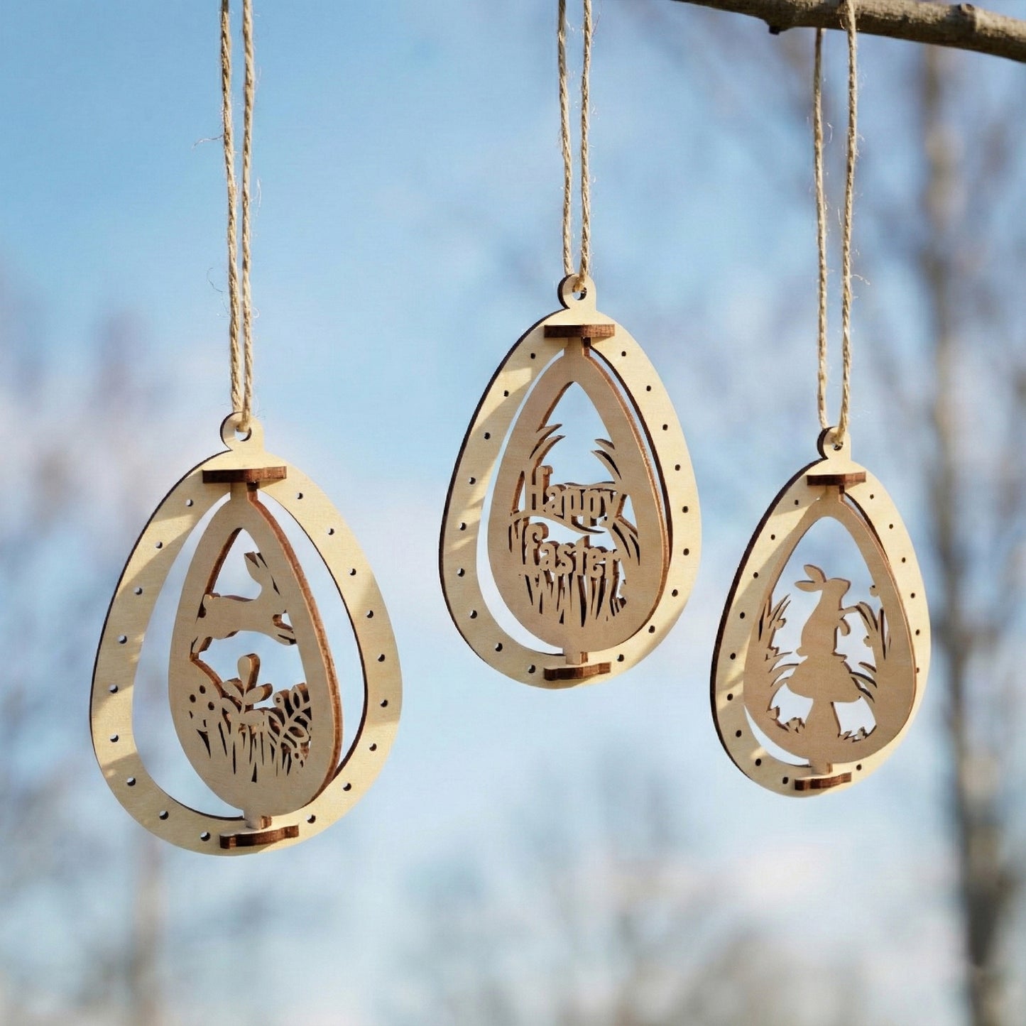 Three laser cut plywood Easter egg ornaments hanging against a blue sky. Each egg features a different cutout: a running rabbit, a "Happy Easter" greeting, and a rabbit by a mushroom. The natural birch wood has elegant dark edges.