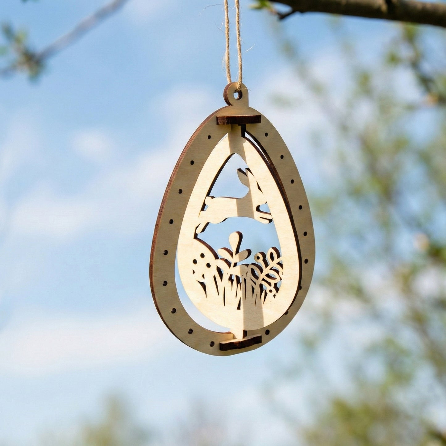 A laser cut plywood Easter egg ornament with a cutout of a rabbit jumping over grass and flowers. The 3D-style wooden frame is suspended by a twine string, showing the smooth wood texture and precise laser-burned details.