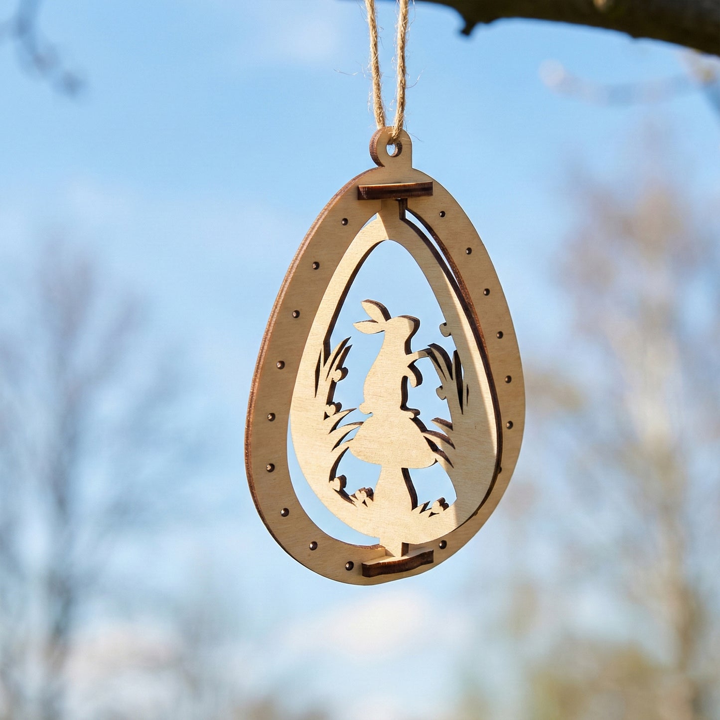 An intricate laser cut plywood egg ornament showing a rabbit standing next to a large mushroom. The delicate wooden silhouette is framed by an outer egg shape with decorative dots, hanging outdoors against a soft bokeh background.