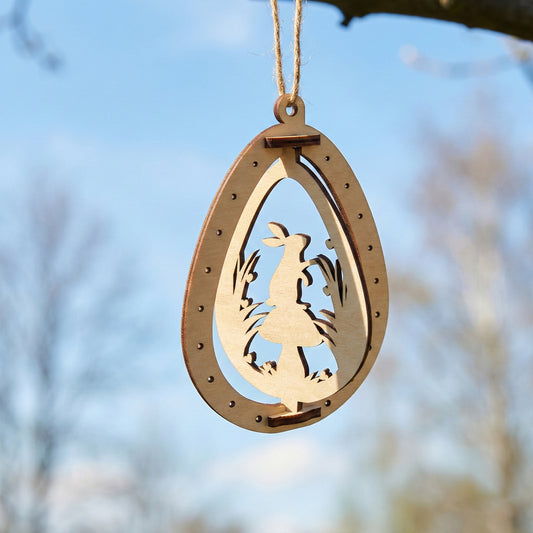 An intricate laser cut plywood egg ornament showing a rabbit standing next to a large mushroom. The delicate wooden silhouette is framed by an outer egg shape with decorative dots, hanging outdoors against a soft bokeh background.
