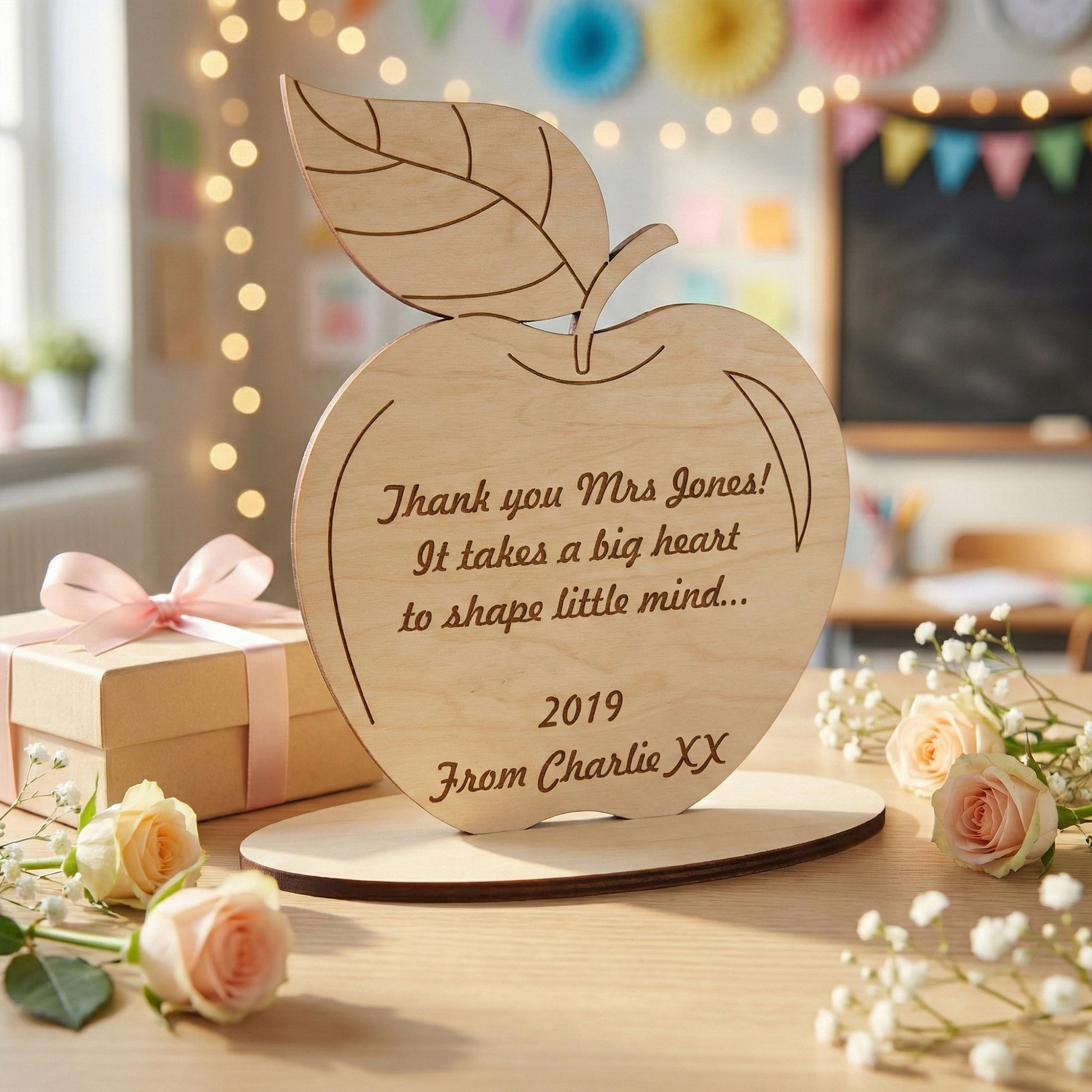 Laser cut plywood plaque in the shape of an apple with a leaf. Engraved with a heartfelt quote for a teacher and personalized from "Charlie XX." The smooth natural wood grain and precise dark edges are shown in a bright classroom.