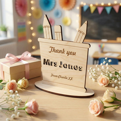 A wooden pencil holder silhouette made of laser cut plywood with three pencils sticking out. Features the teacher's name "Mrs Jones" in bold cutout letters. Set on a wooden table alongside a gift and roses for a teacher's thank you gift.