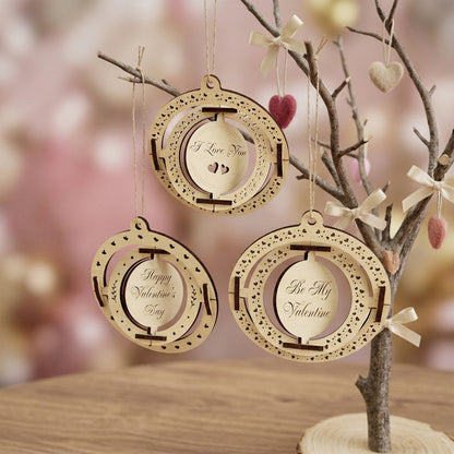 A set of three kinetic Valentine's Day ornaments made from laser-cut plywood. Each ornament features nested spinning rings and engraved messages: "I Love You," "Happy Valentine's Day," and "Be My Valentine." Hanging from a decorative branch in a soft, romantic setting with warm lighting.