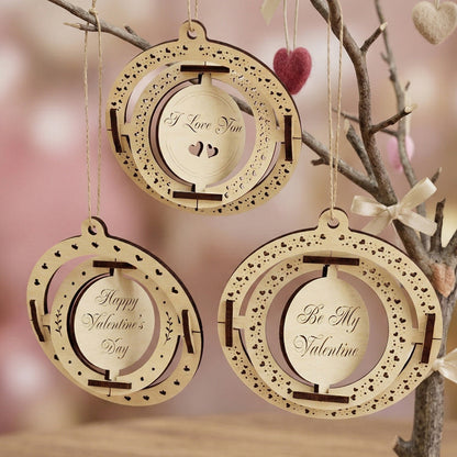 A set of three kinetic Valentine's Day ornaments made from laser-cut plywood. Each ornament features nested spinning rings and engraved messages: "I Love You," "Happy Valentine's Day," and "Be My Valentine." Hanging from a decorative branch in a soft, romantic setting with warm lighting.