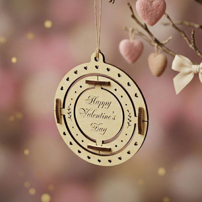 Close-up of a 3D spinning laser-cut wooden Valentine's card with "Happy Valentine's Day" engraved in the center. The design features an outer ring with heart cutouts and inner rotating parts. Posed hanging from a branch against a blurred, aesthetic pink background.