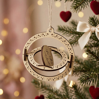 Side-angled view of a rotating 3D wooden Valentine's Day ornament with "Be My Valentine" engraving. This kinetic gift highlights the nested spinning rings and heart-patterned borders. Displayed hanging on a pine branch with warm festive lights in the background.