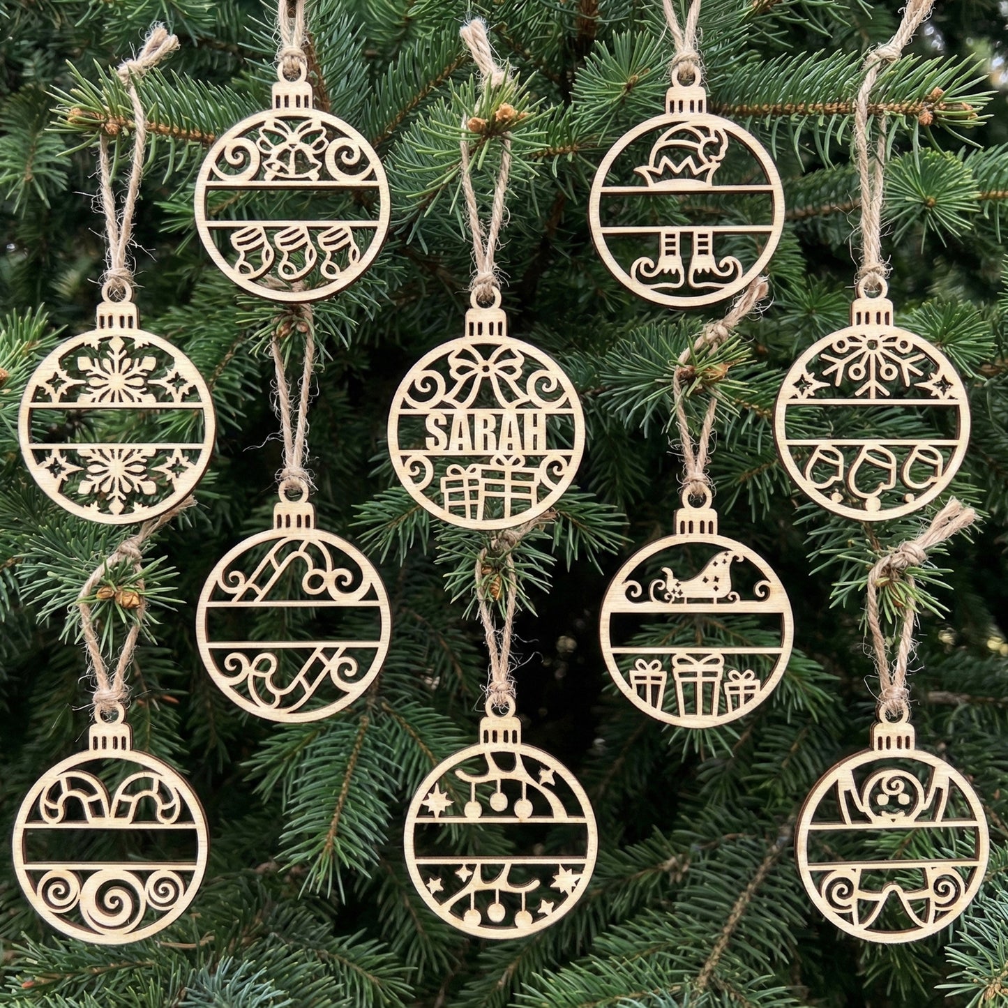 Collection of wooden laser cut Name Christmas ornaments in bauble shapes with personalized split monogram designs, festive motifs like snowflakes and reindeer, hanging on a natural green Christmas tree background.
