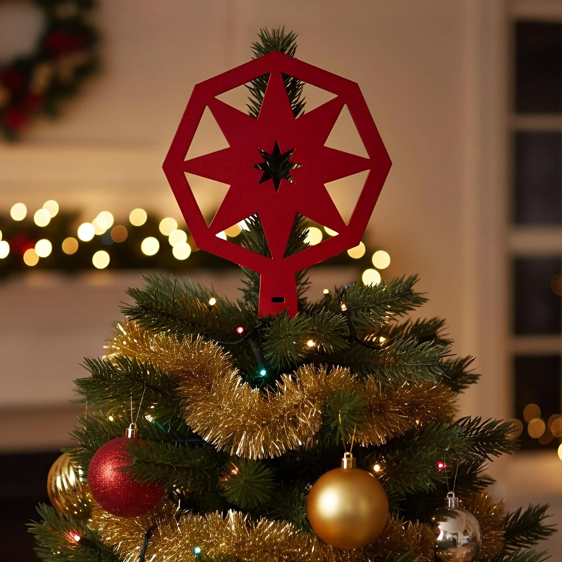Modern laser cut wooden tree topper in bold red with intricate star and snowflake cutouts, displayed on a lit Christmas tree against a softly blurred festive background.
