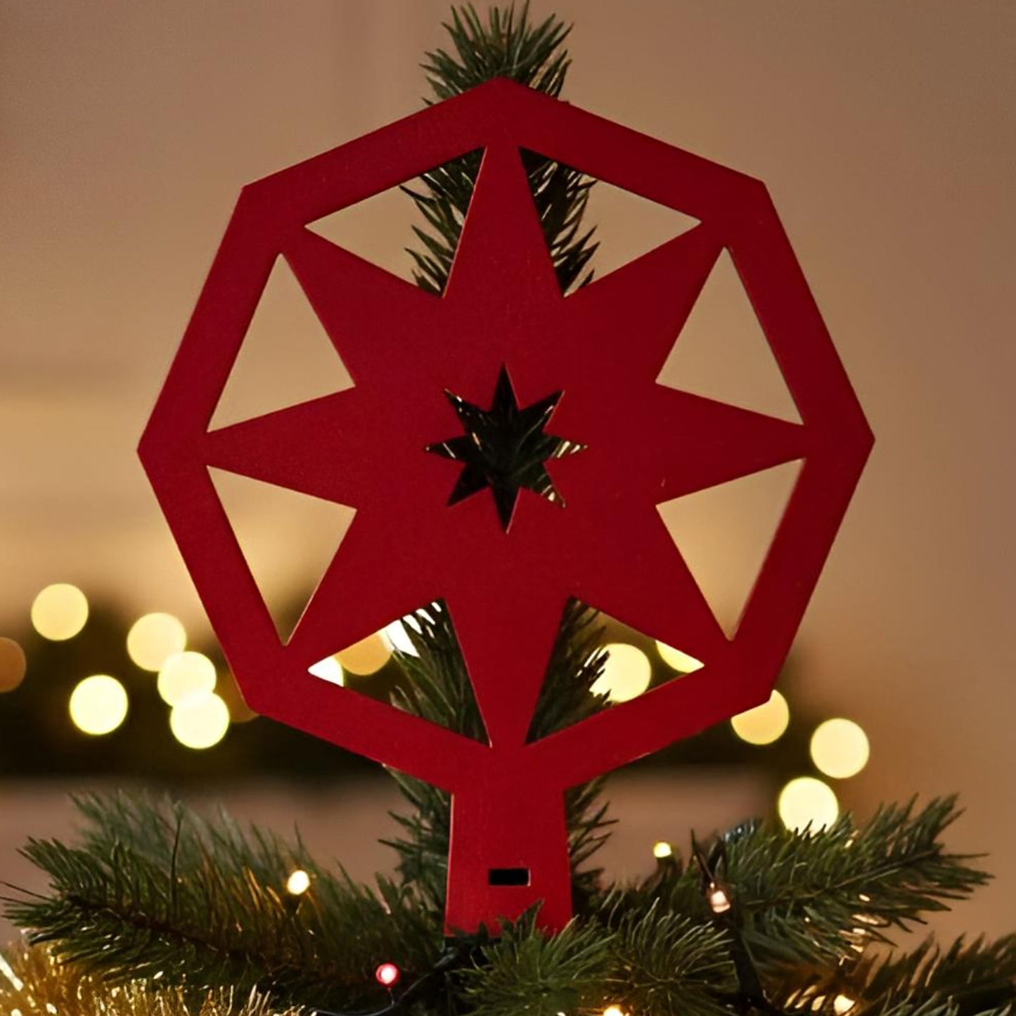 Red wooden laser cut Christmas tree topper featuring a geometric snowflake-star design, shown atop a decorated Christmas tree with warm bokeh lights in the background.