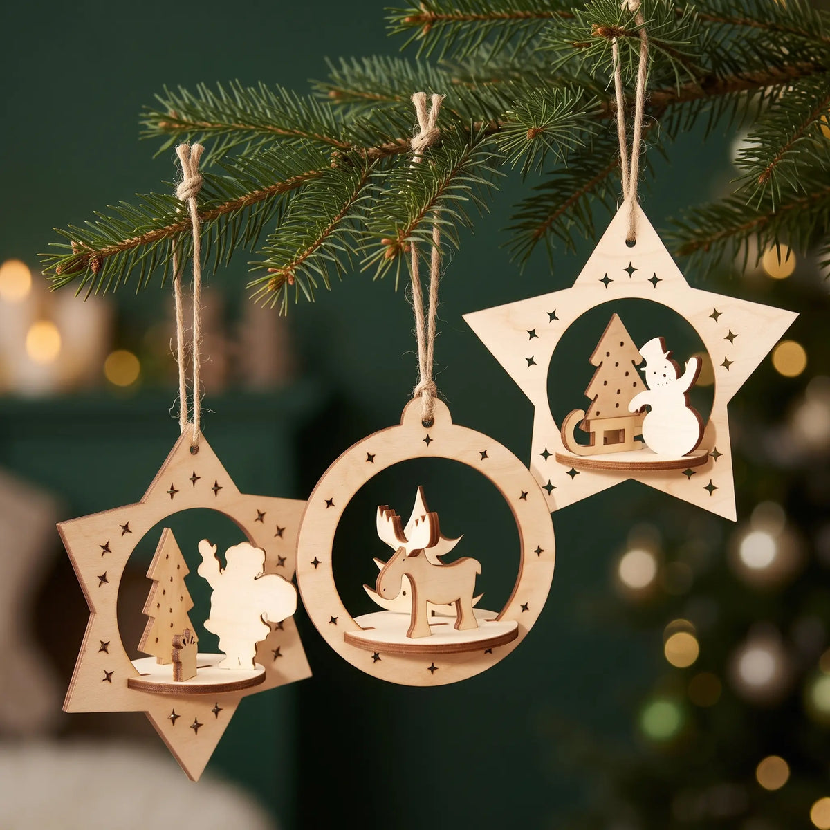 Three laser cut plywood 3D ornaments with deer, snowman, and Santa hanging from pine branch with lights.