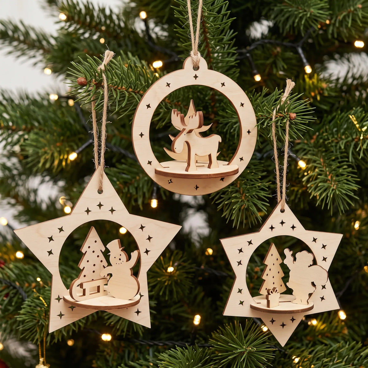 Trio of laser cut plywood holiday baubles with miniature dioramas hanging on lush Christmas pine tree.