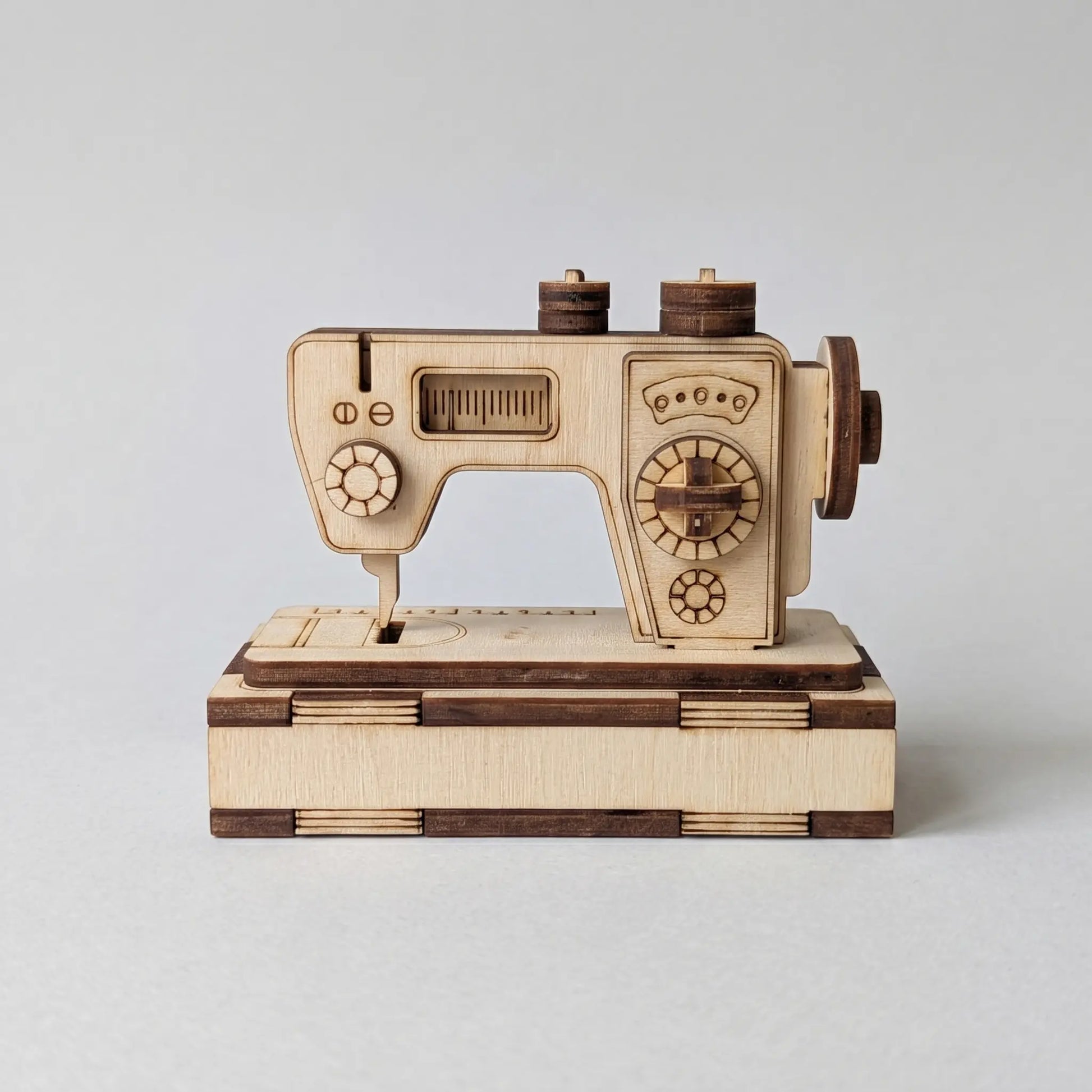 A miniature laser cut sewing machine model with a light natural wood body and dark brown accents, displayed on a matching wooden base against a neutral light gray background.