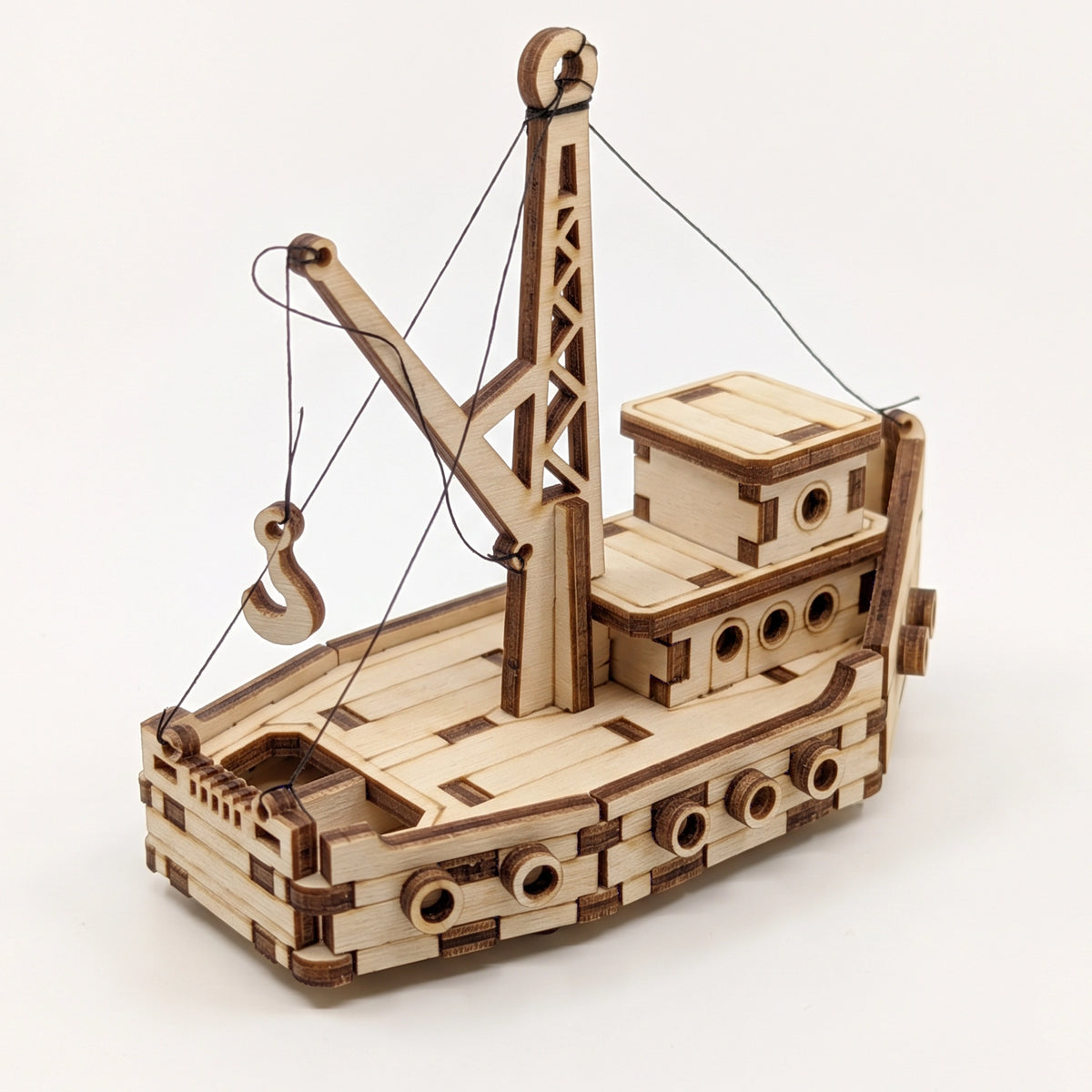 High angle view of 3D laser cut plywood crane boat model with rigging, hook, and round portholes on plain white background.
