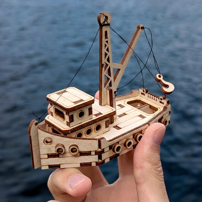 Hand holding miniature laser cut plywood tugboat model with boom crane and thread rigging above rippling ocean waves.