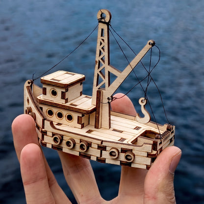 Hand holding assembled 3D laser cut plywood crane vessel model showing scale over deep blue rippling water background.